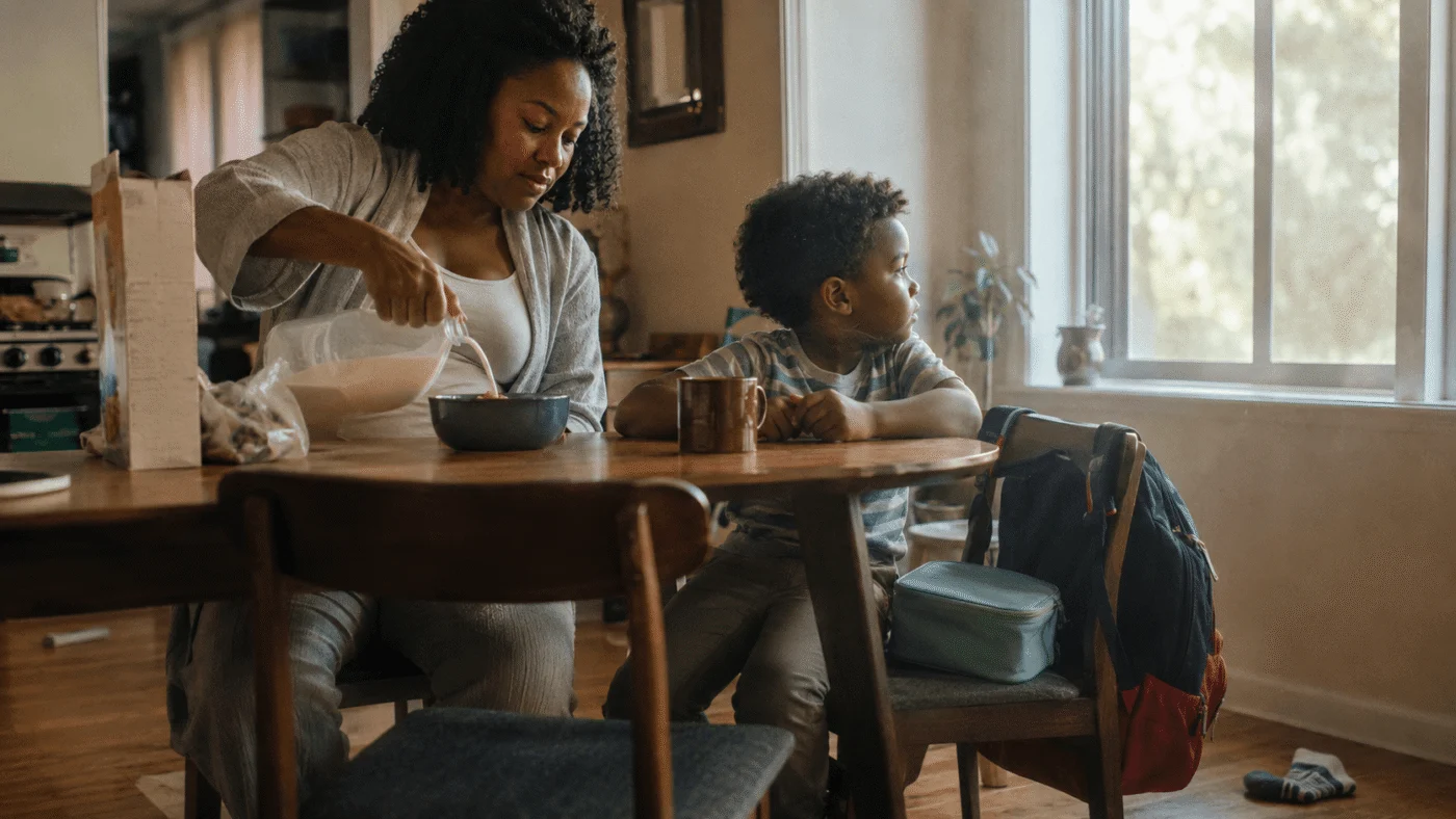 A mother pours breakfast cereal at a sunny kitchen table beside her young son, his packed school backpack on the chair
