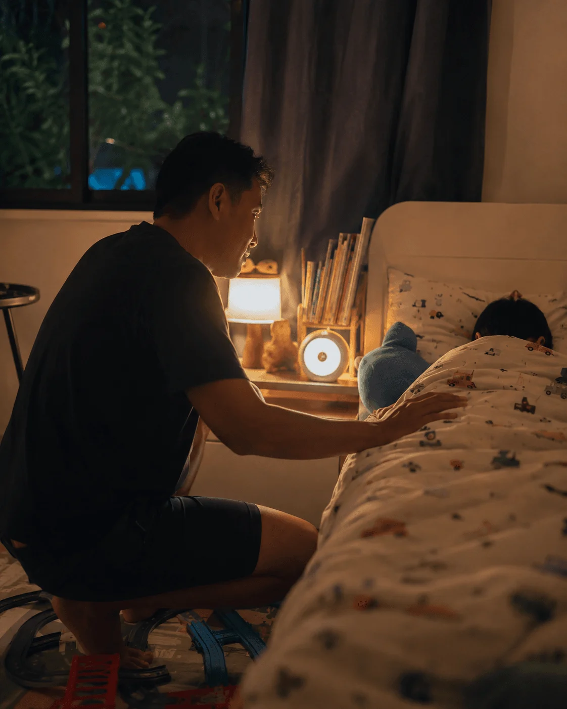 A father kneels by his child's bed at dusk, a visual timer with a soft night light glowing on the nightstand during the bedtime wind-down