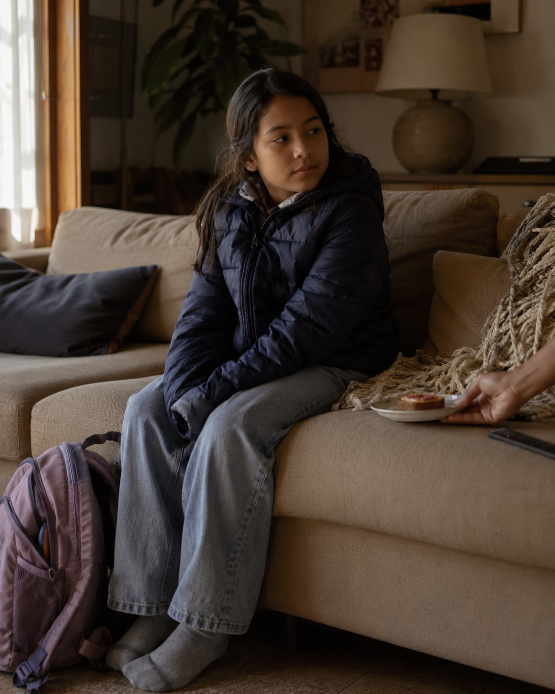 A child sits quietly on a sofa with a backpack after arriving at a parent's home, someone offering her a snack