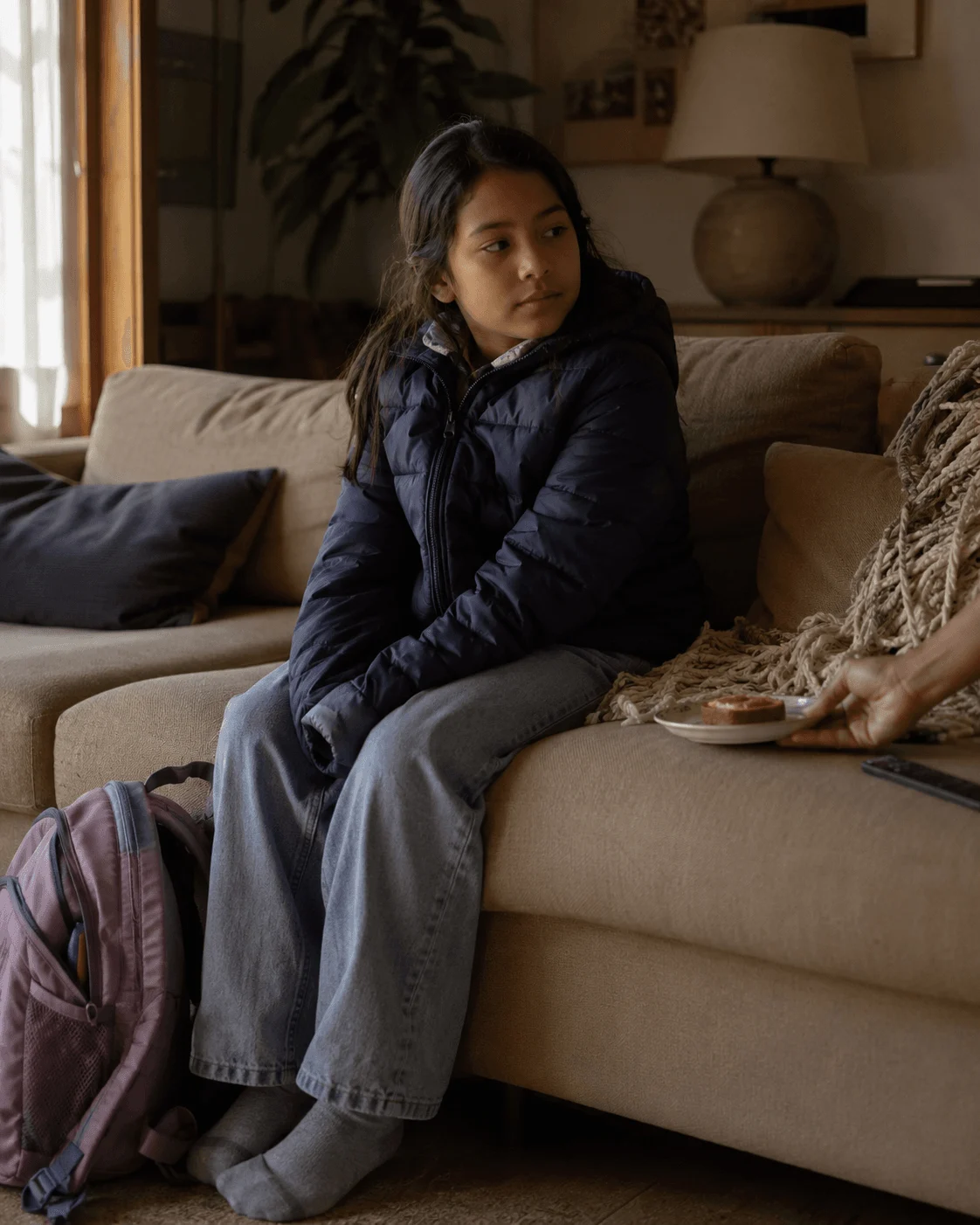 A child sits quietly on a sofa with a backpack after arriving at a parent's home, someone offering her a snack