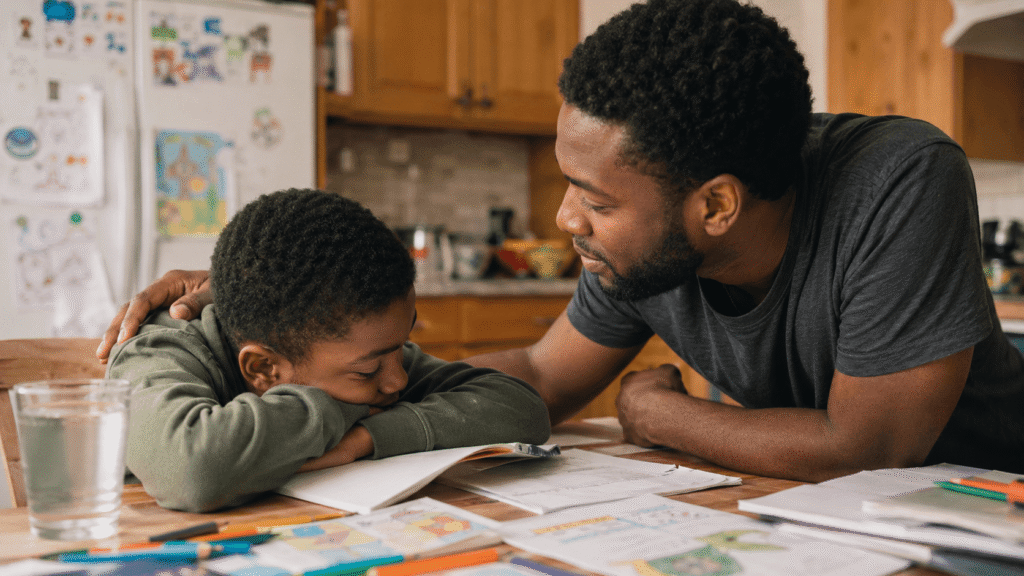 A father kneels beside his young son at a homework-covered kitchen table with a gentle hand on his back, a calm and connected moment