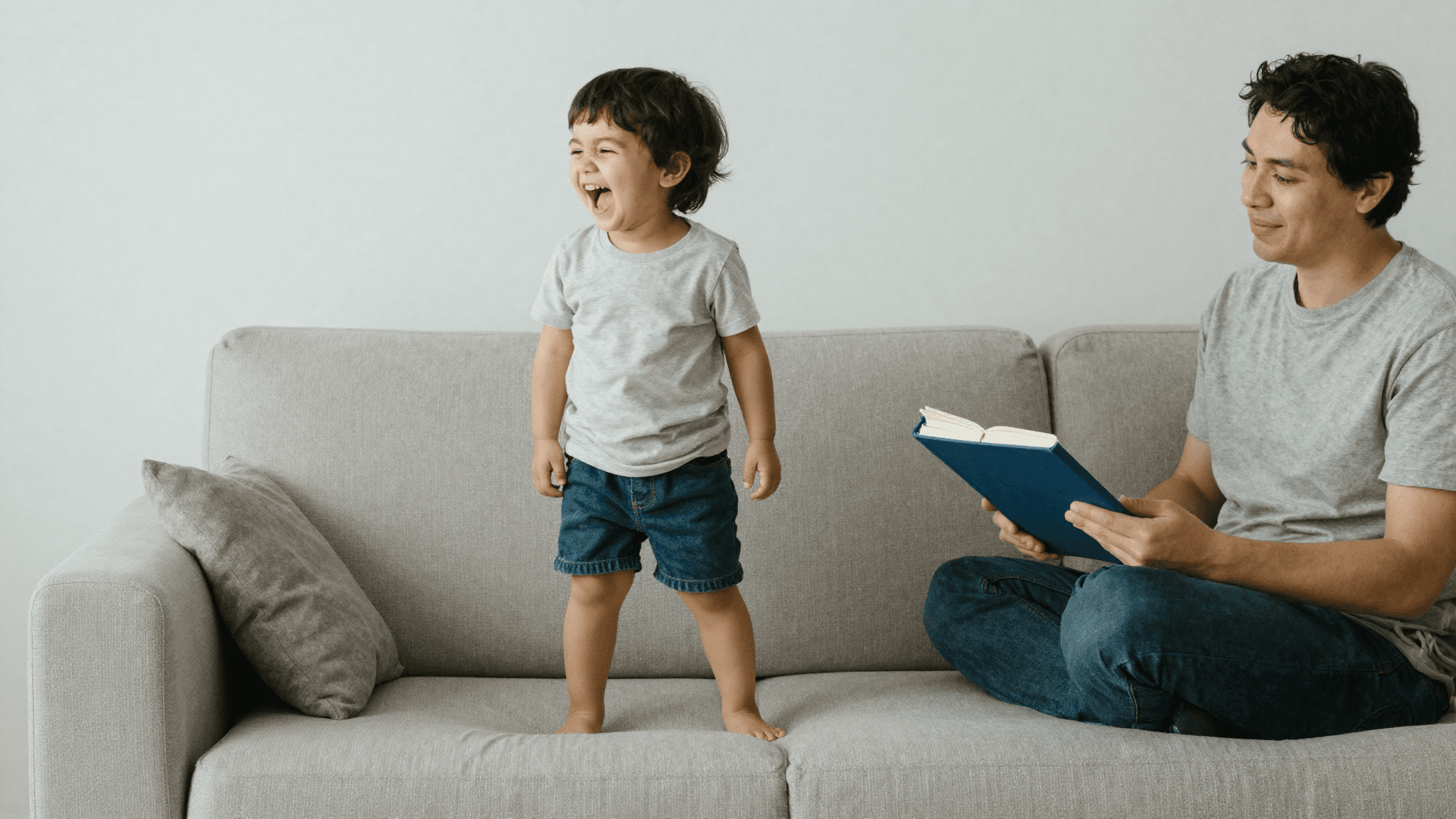 A calm parent watching a spirited young child laughing on the living-room sofa