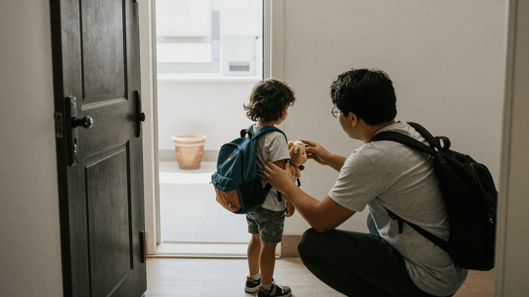A child with a small backpack and soft toy at the front door as a parent crouches to say goodbye