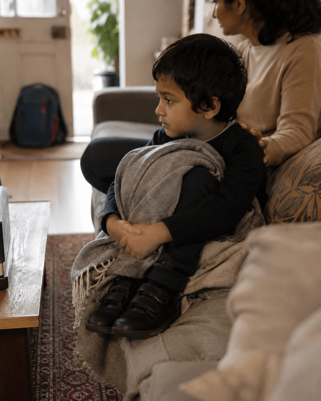 A young child sits tucked up on the sofa under a blanket with school shoes on, a packed backpack nearby and a parent resting calmly at the other end
