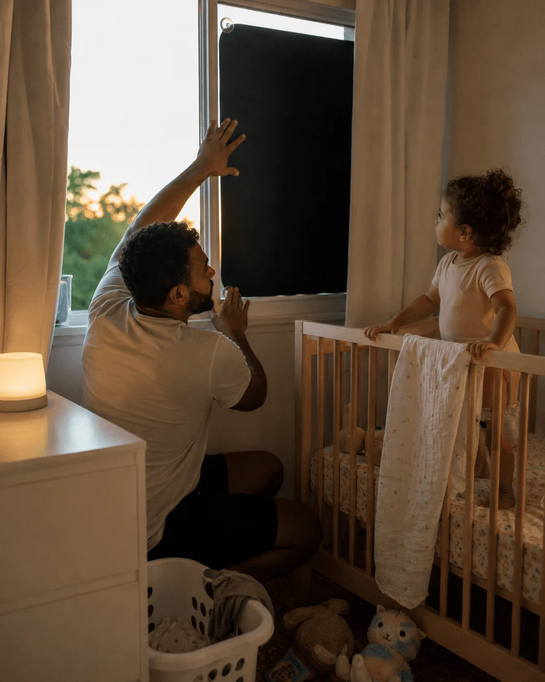A young father presses a portable blackout blind onto the window as his toddler watches from the cot, the summer evening light half-darkened