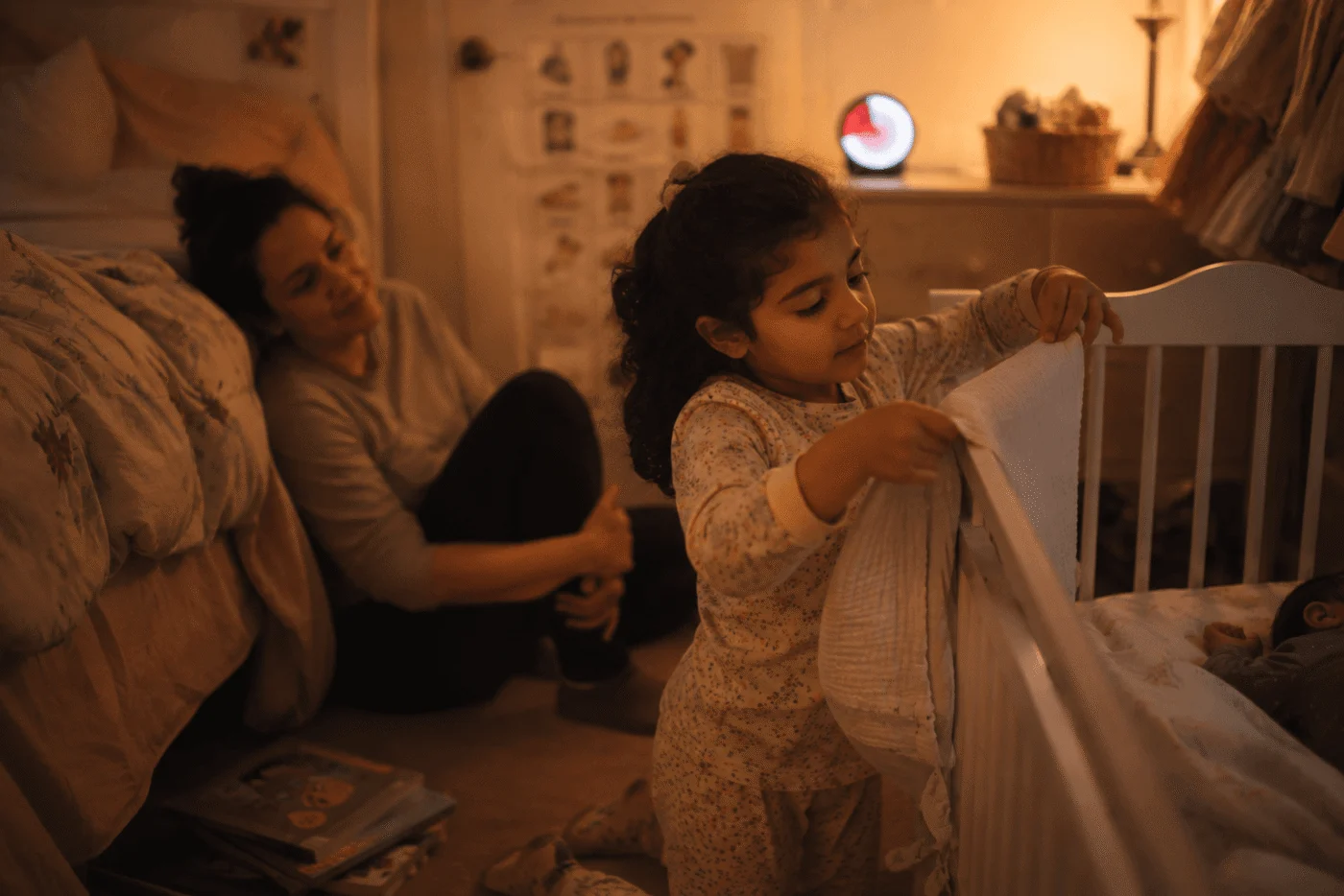 A small girl kneels proudly by a baby's cot laying a folded muslin over the rail as her helper job, her tired mother watching from the floor, a visual timer glowing nearby