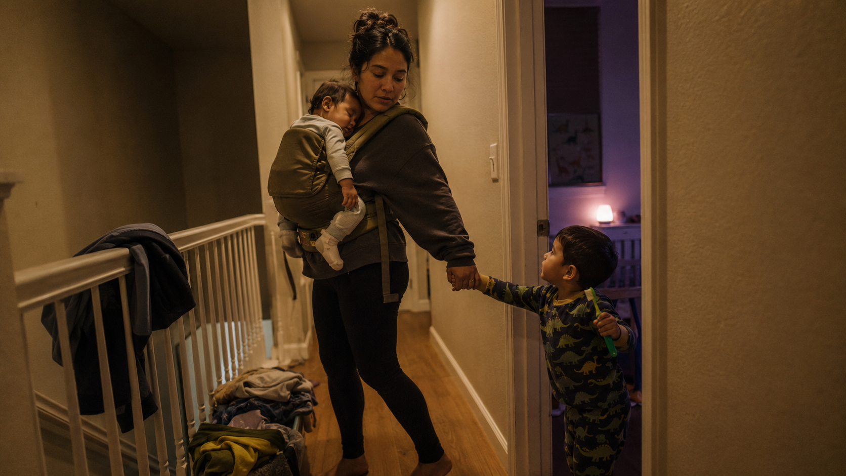A young single mother stands on an upstairs landing with a drowsy baby worn on her chest and her toddler in pyjamas tugging her free hand toward the bedroom