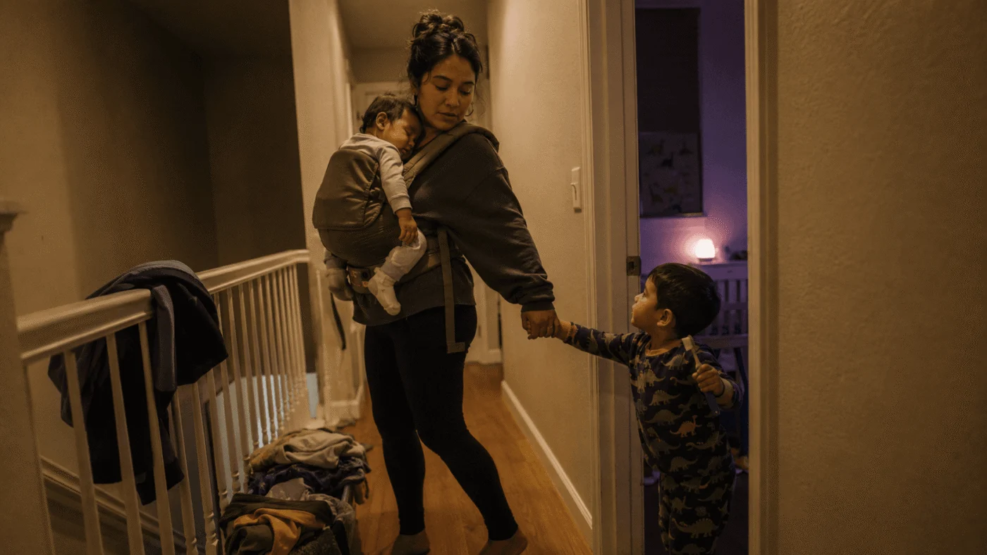 A young single mother stands on an upstairs landing with a drowsy baby worn on her chest and her toddler in pyjamas tugging her free hand toward the bedroom