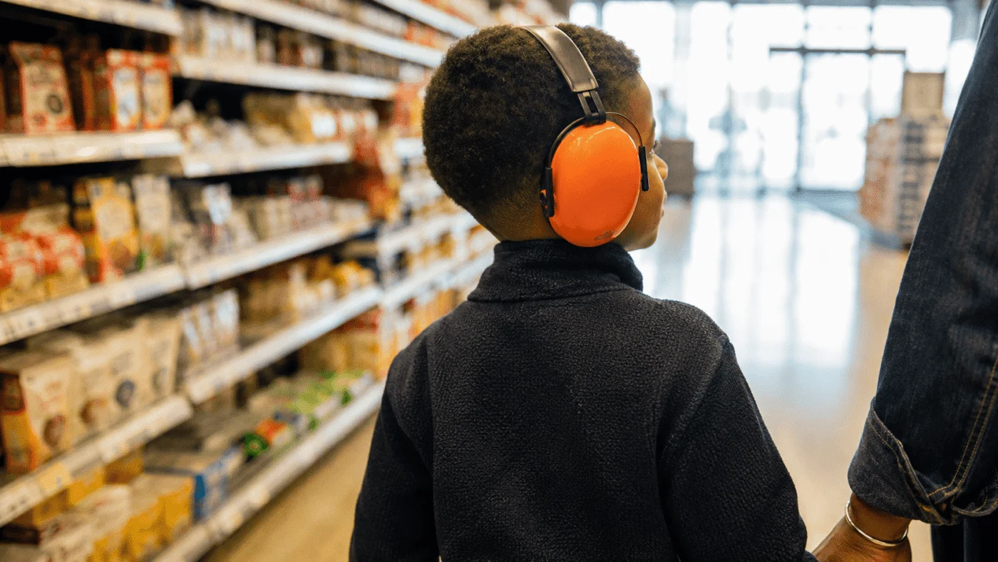 A Black boy wearing orange ear defenders walks calmly through a supermarket aisle, seen from behind with a parent beside him