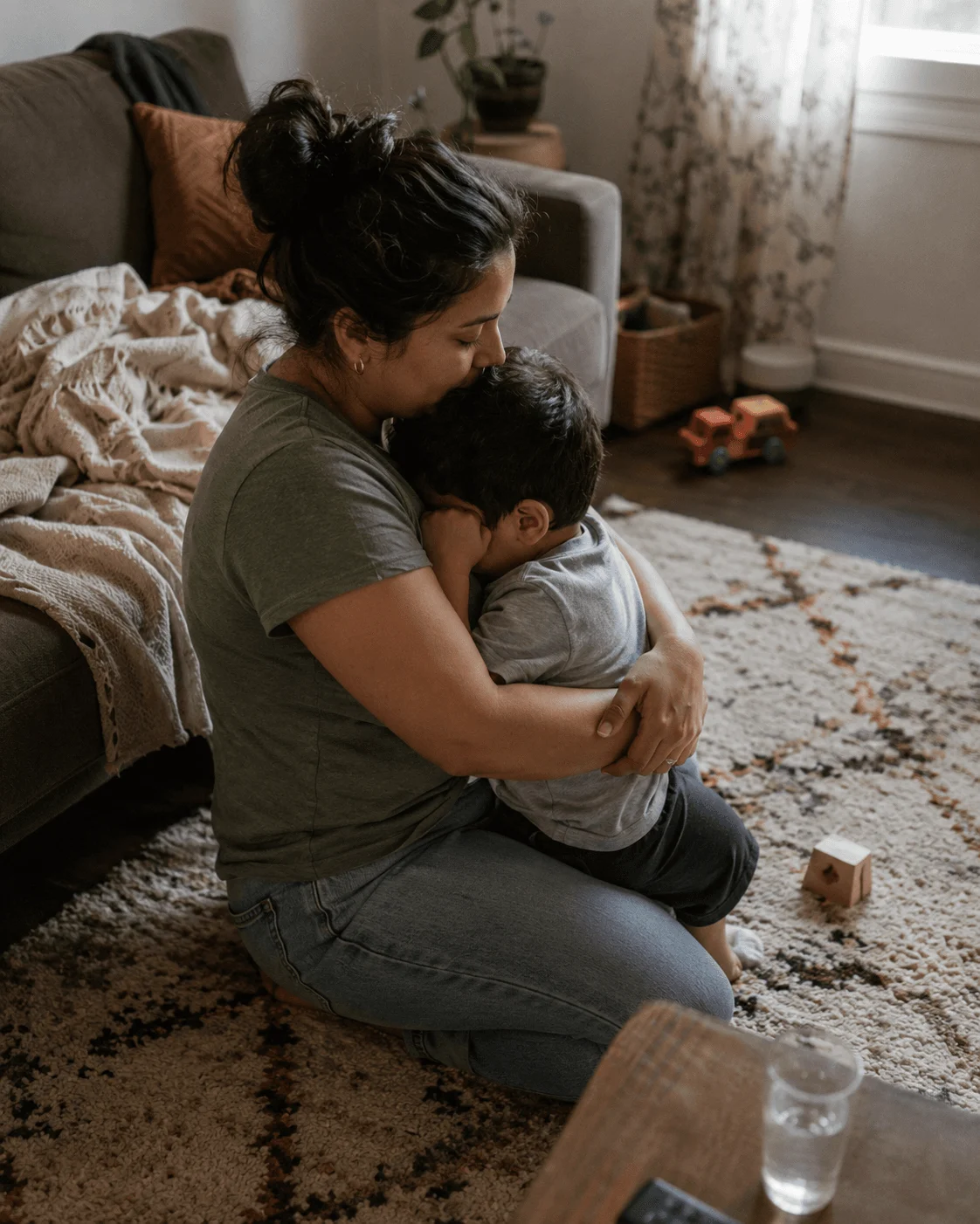 A Latina mother sits on the living-room floor holding her young son in a calm, close hug, his face resting against her chest, toys and soft daylight around them