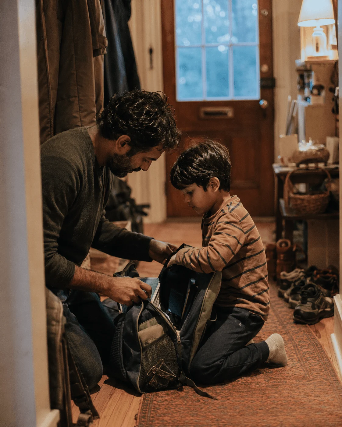 A father kneels with his young son in a warm hallway, the two of them packing a school backpack together the night before
