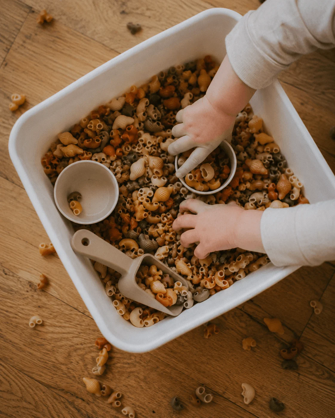 Overhead view of a toddler's hands scooping colourful dried pasta in a white sensory bin on a wooden floor