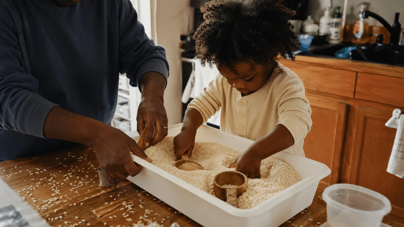 A father and his young son run their hands through a tray of dried rice with small scoops at a kitchen table in warm daylight