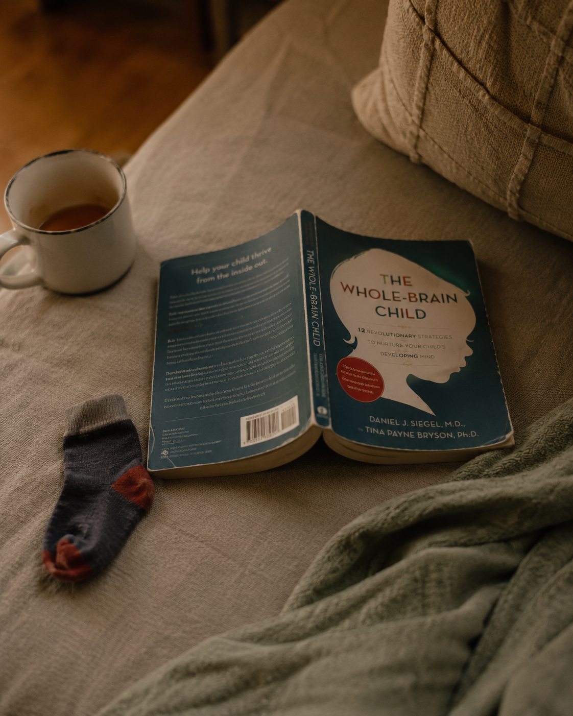 A copy of The Whole-Brain Child by Dan Siegel and Tina Payne Bryson lies open on a linen sofa beside a mug of tea and a child's sock