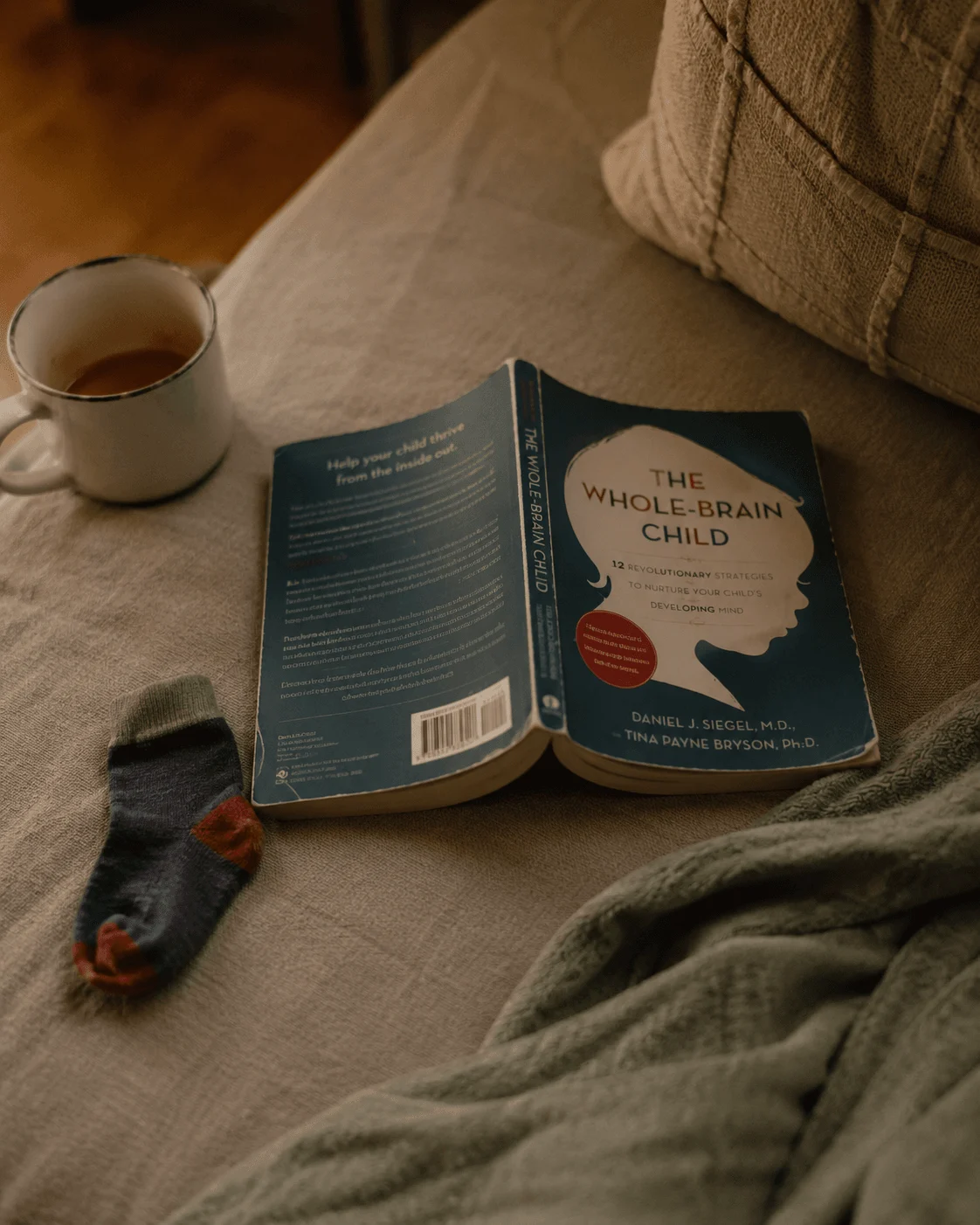 A copy of The Whole-Brain Child by Dan Siegel and Tina Payne Bryson lies open on a linen sofa beside a mug of tea and a child's sock