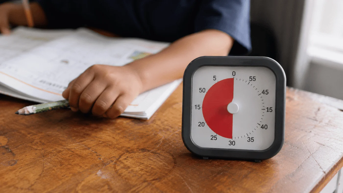 A Time Timer MOD counting down on a wooden desk, a child's hand and an open workbook softly out of focus behind it