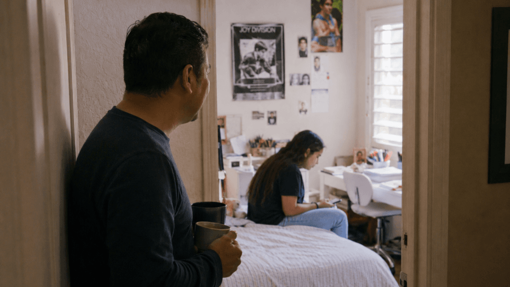 A father stands in a hallway looking into his teenage daughter's bedroom in the evening, watching her sit on the bed with her phone near an unfinished homework desk