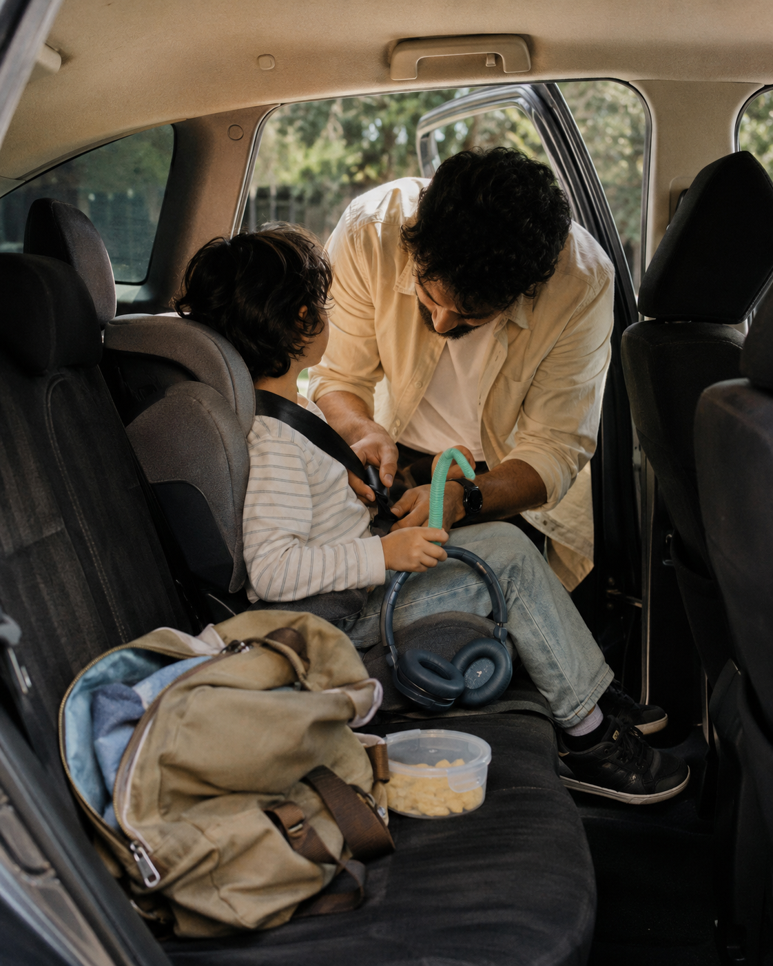 A Middle Eastern father leans into the car buckling his young son into his car seat, blue ear defenders and a chew toy ready on the seat beside the snack box