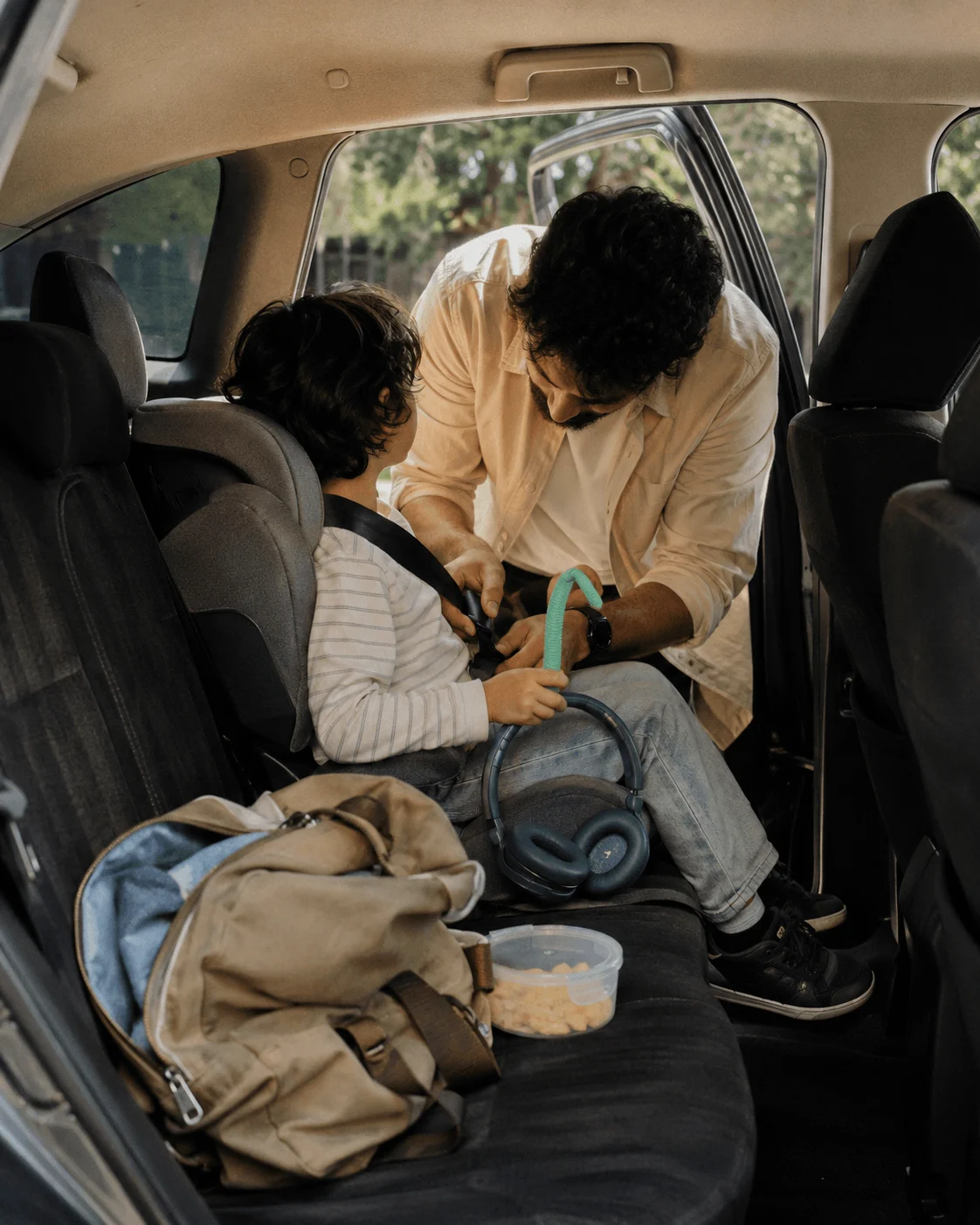 A Middle Eastern father leans into the car buckling his young son into his car seat, blue ear defenders and a chew toy ready on the seat beside the snack box