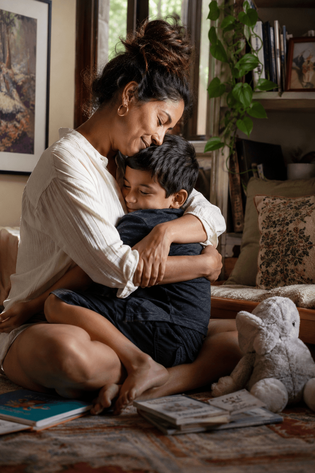 A South Asian mother sits cross-legged on the living-room floor hugging her young son tightly, eyes closed, picture books and a stuffed elephant on the rug beside them