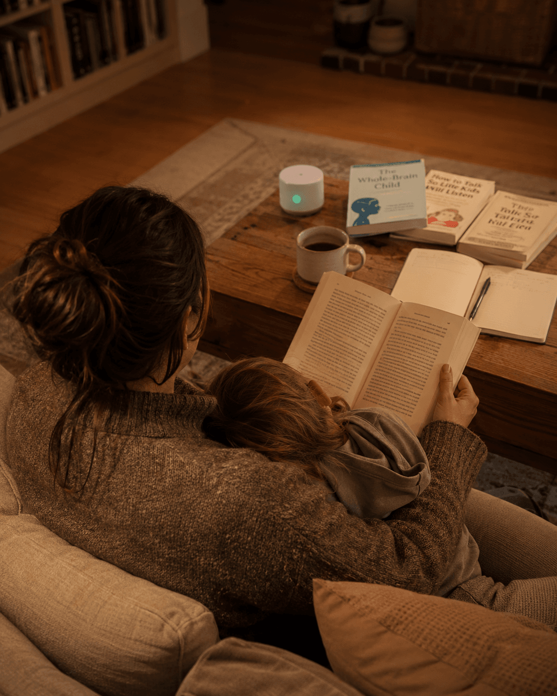 A mother reads on the sofa with her child snuggled against her, several parenting books laid out on the coffee table in warm evening light
