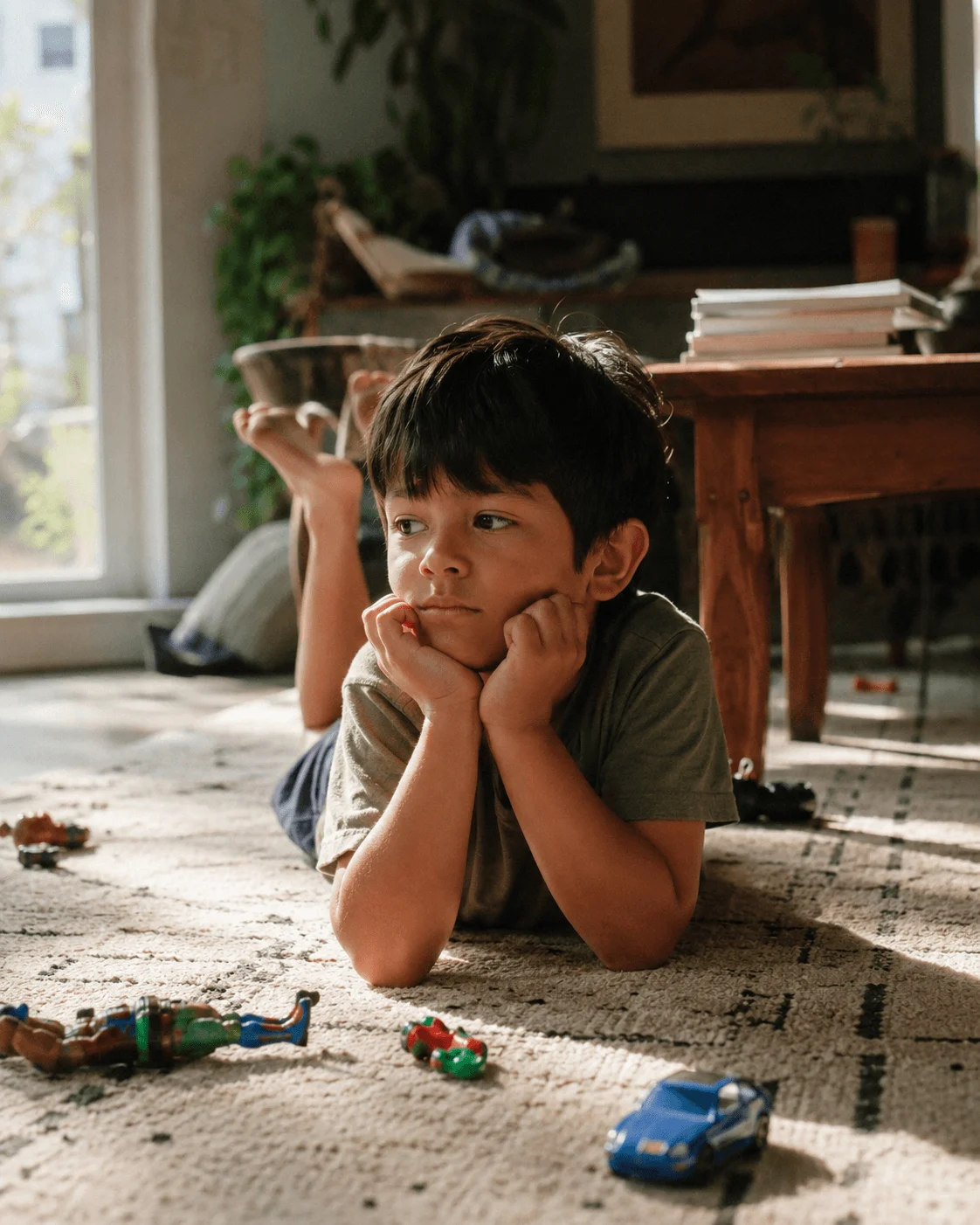 A Latino boy lies on the living-room rug, chin on his hands, gazing idly, toy cars untouched nearby