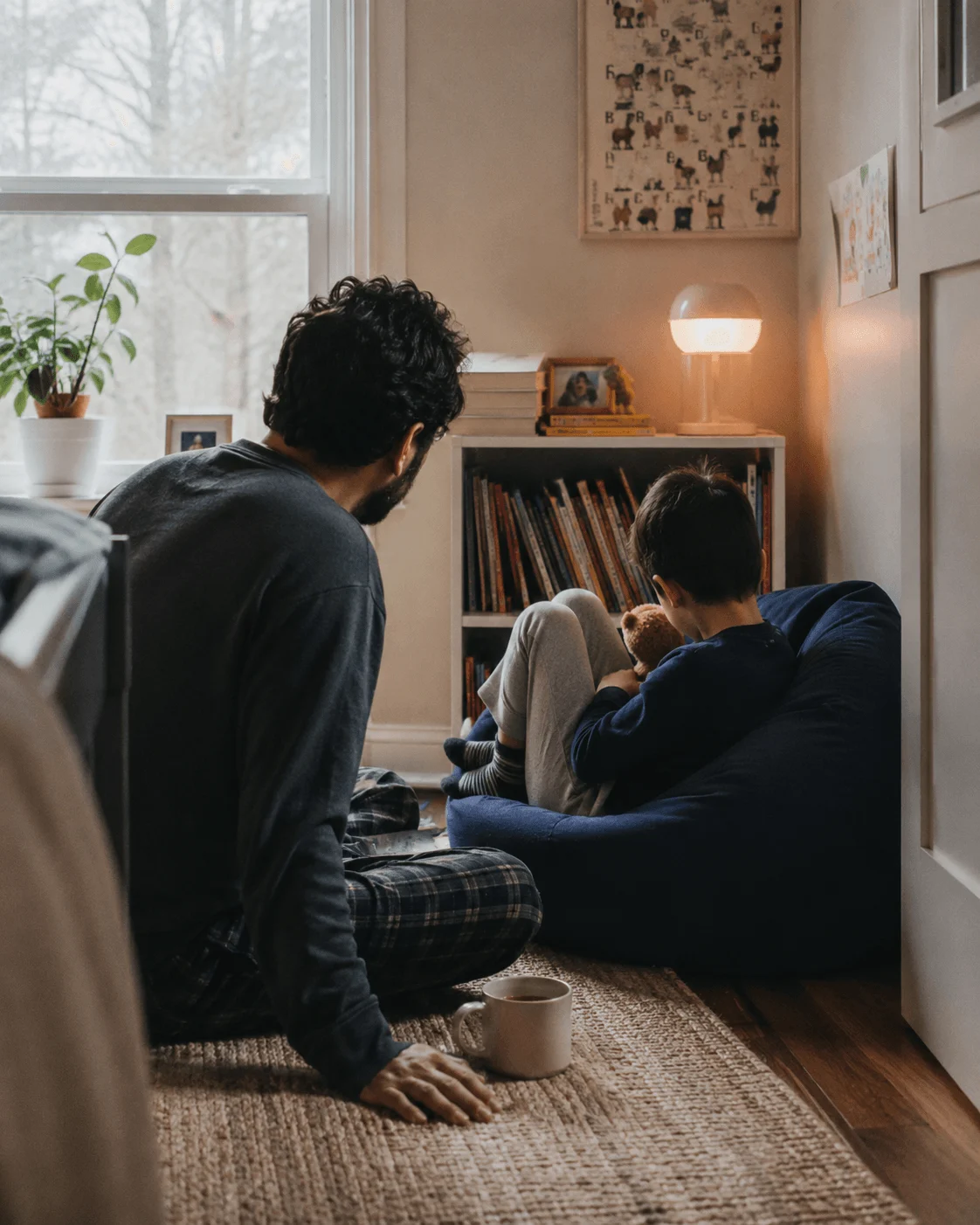 A Middle Eastern father and his young son sit close together in a cozy reading corner, a small lamp lit beside a bookshelf