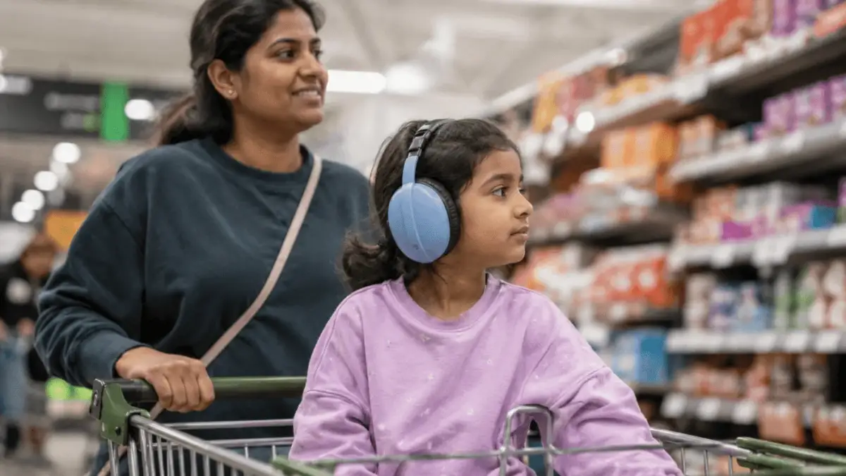 Girl wearing light blue headphones sitting in a shopping cart while her mother pushes it through a supermarket