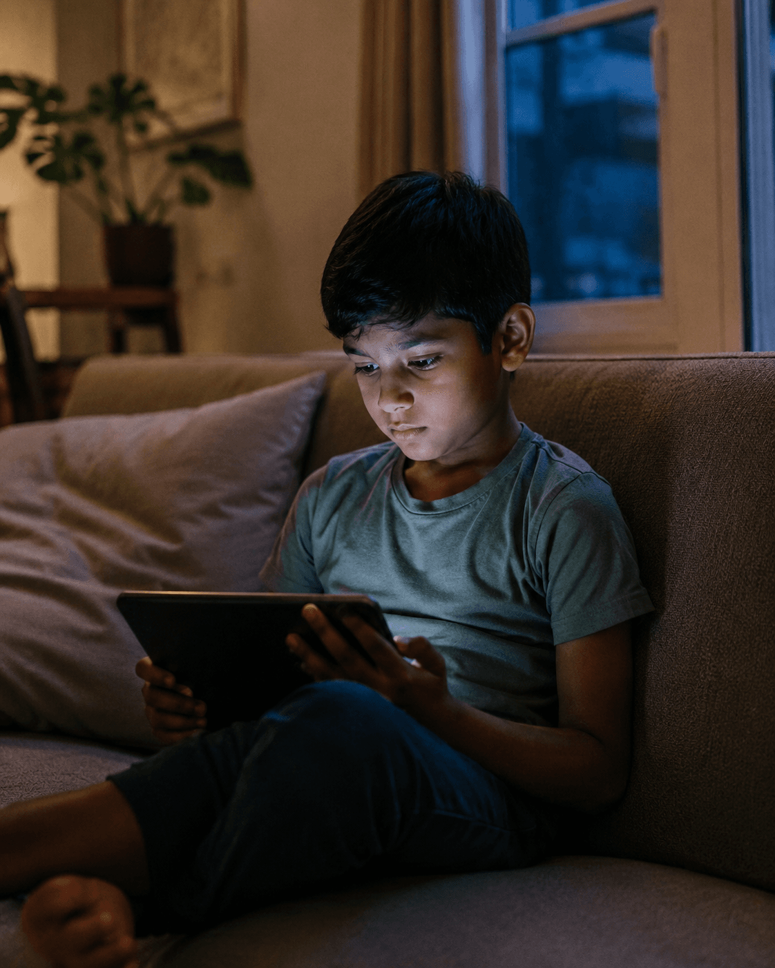 A South Asian boy sits alone on a couch in the evening, his face lit only by the tablet in his hands
