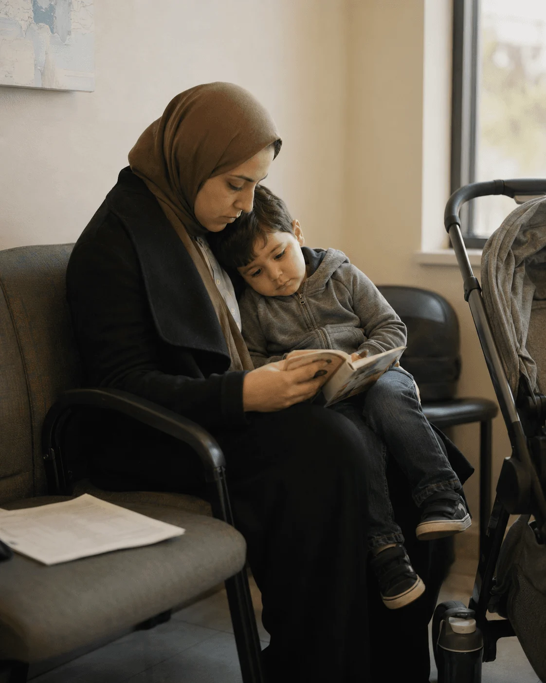 A mother in a hijab reads a picture book with her son on her lap in a clinic waiting room, intake paperwork on the chair beside her
