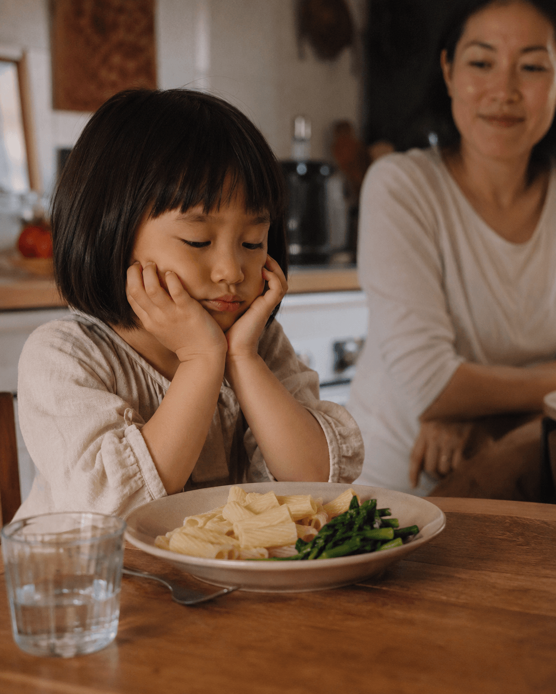 A young East Asian child looks at a dinner plate of pasta and asparagus, a relaxed parent in the background