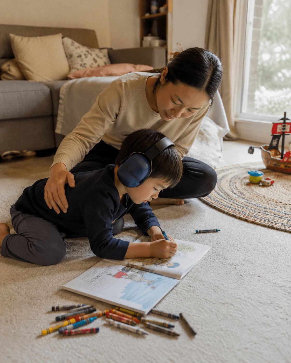 Mother and toddler on a living room floor, the child in navy ear defenders colouring with crayons