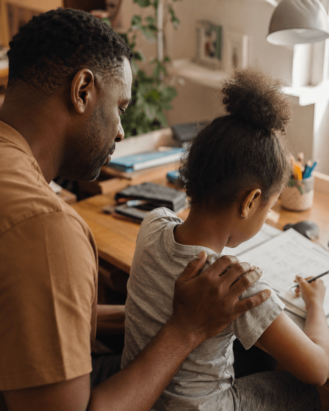 A parent rests a hand on their child's shoulder at a homework-covered table, both looking down at the same page in soft window light