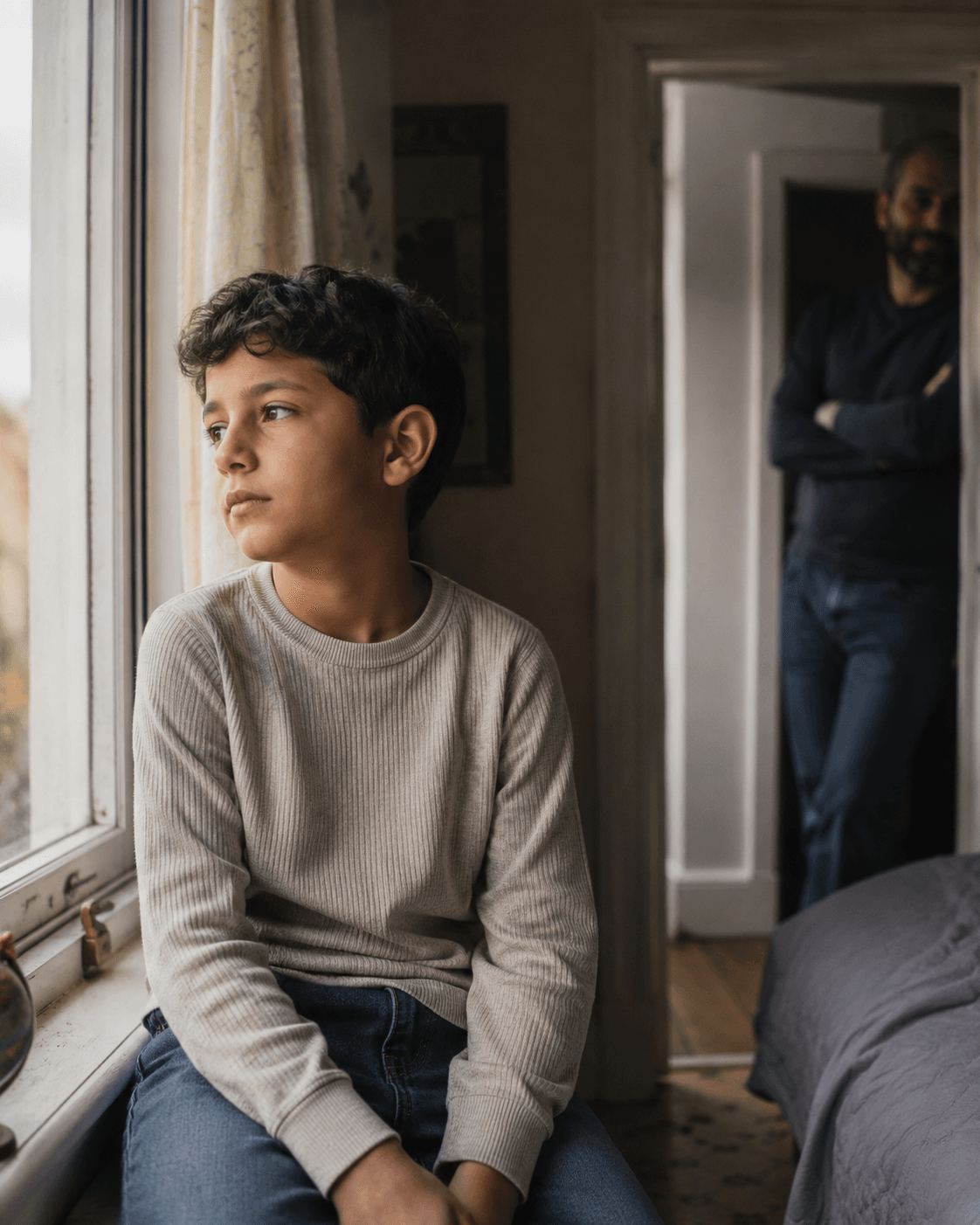 A Middle Eastern boy sits on a windowsill looking out thoughtfully, a parent in the doorway giving him space