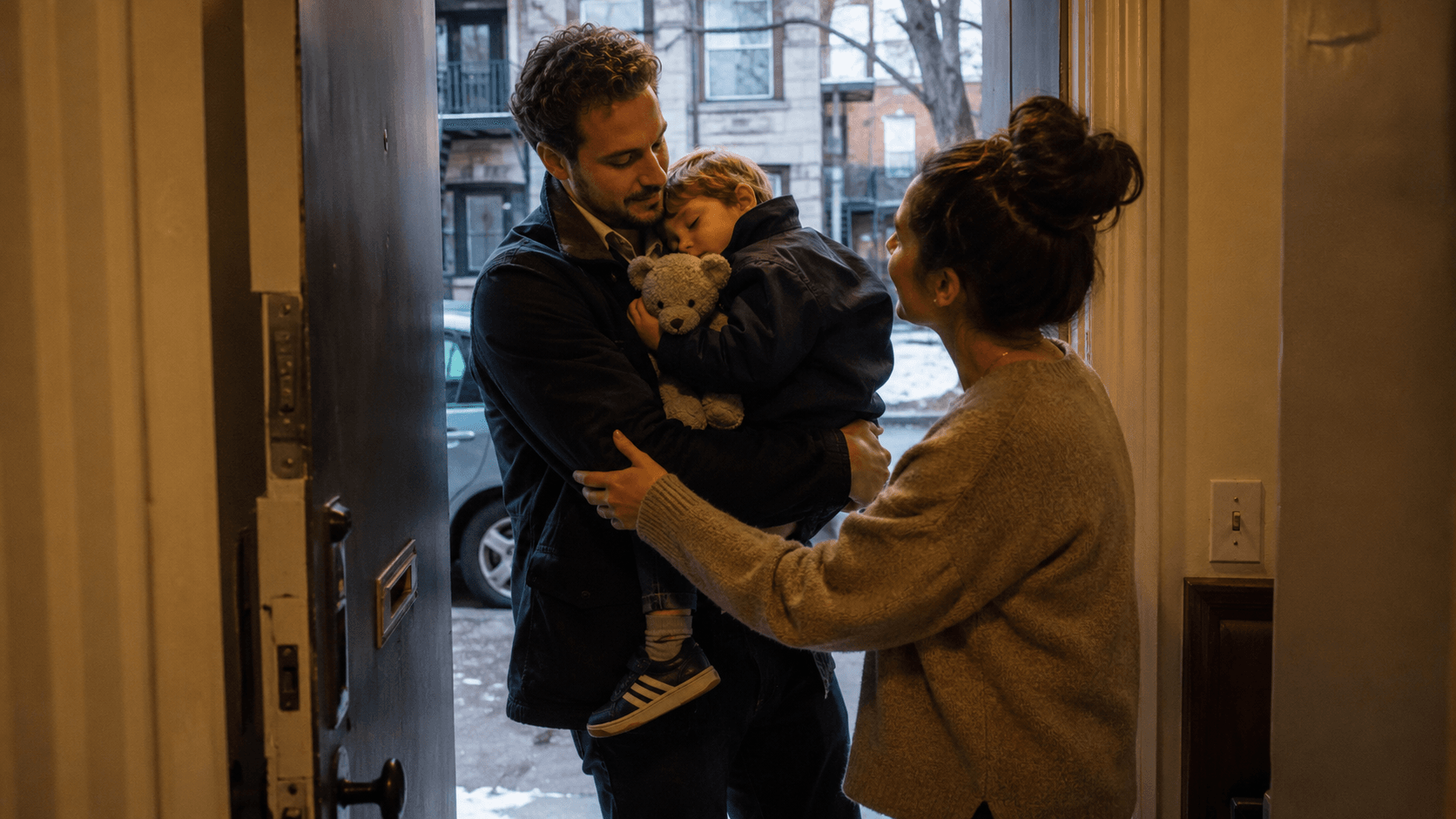 A father stands in a doorway at night holding his sleeping son, handing him over to the boy's mother who reaches out to take him