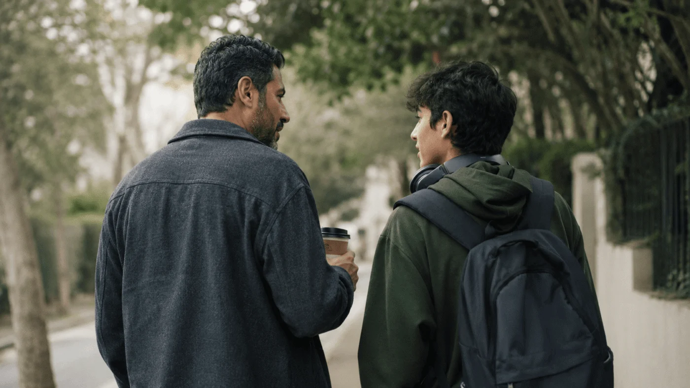 A Middle Eastern father and his teenage son walk down a tree-lined street in quiet conversation