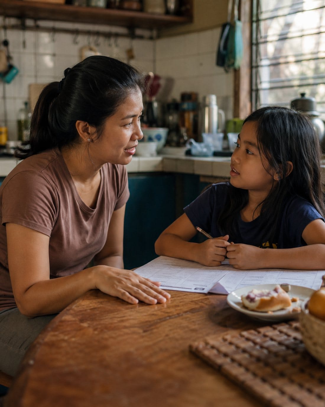A mother crouched at eye level beside her young daughter at a kitchen table, the two of them talking over a homework sheet