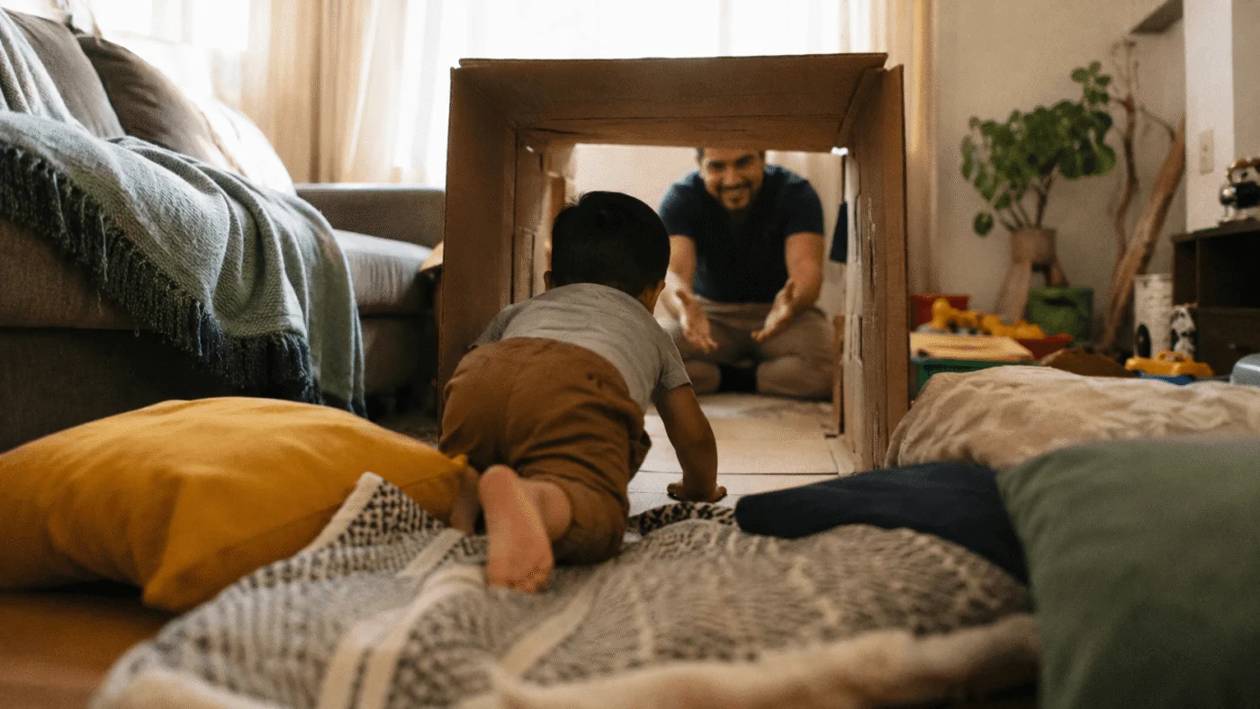 A father and baby crawl through a cardboard box tunnel surrounded by pillows in the living room