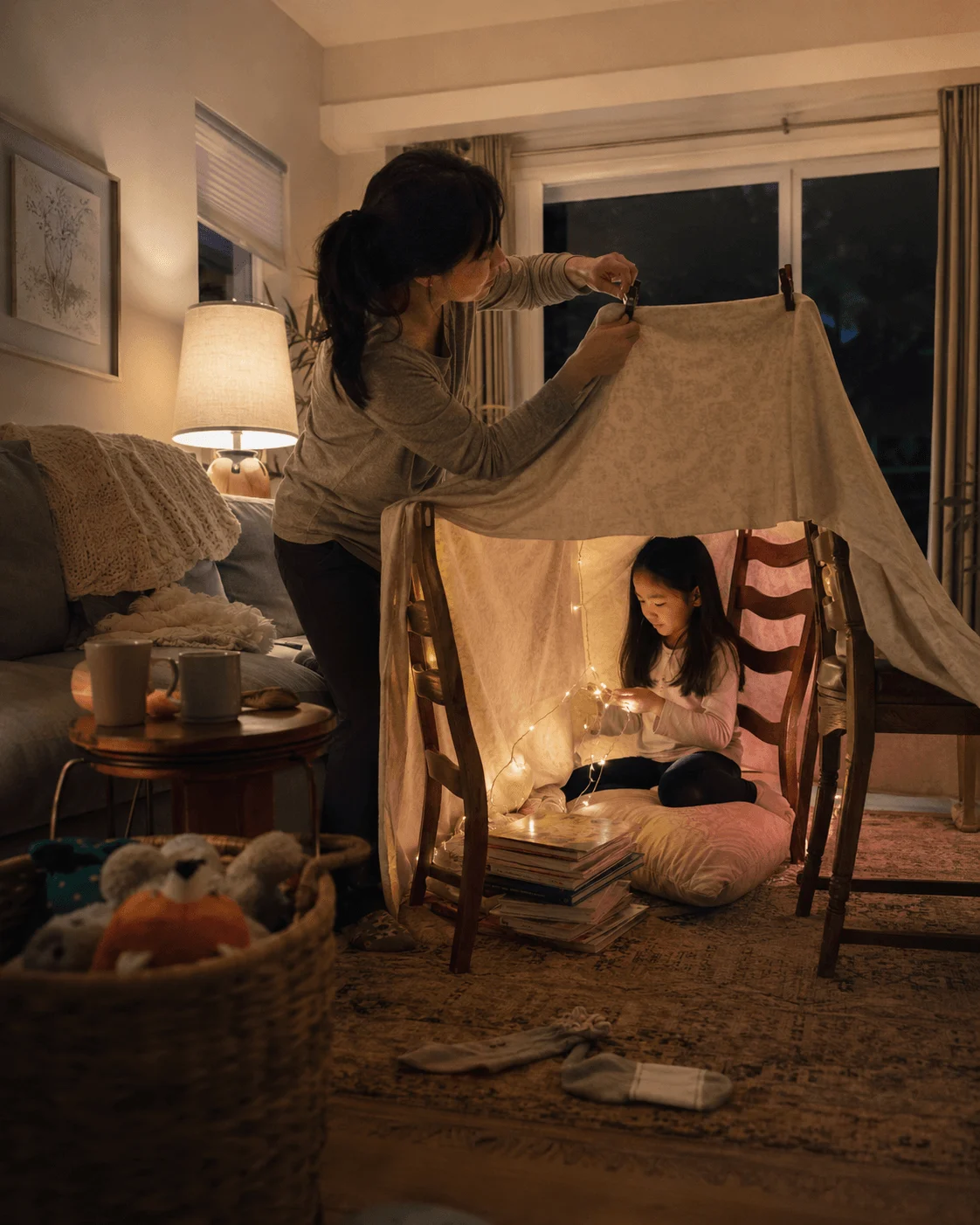 A mother and daughter sit inside a blanket fort with fairy lights and books at night