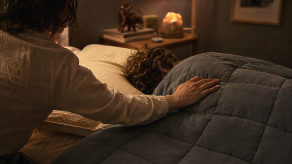 A child tucked into bed under a soft weighted blanket while a parent smooths the corner