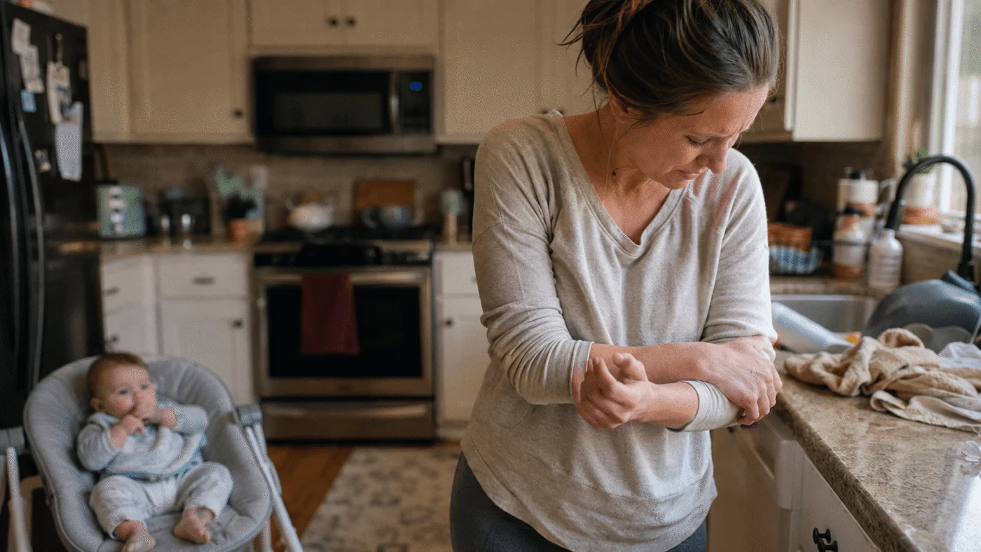 A parent rubs a sore wrist and forearm at the kitchen counter while a baby sits nearby, an ordinary everyday moment of parenting strain