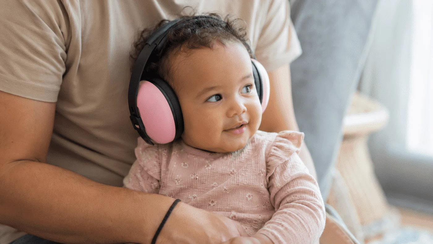 Baby wearing oversized pink ear defenders sits contentedly on a parent's lap