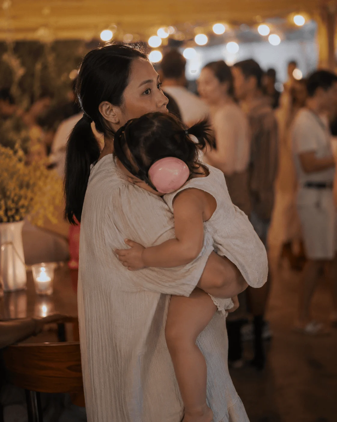 Mother carries her toddler wearing pink ear defenders through a busy, warmly lit indoor event