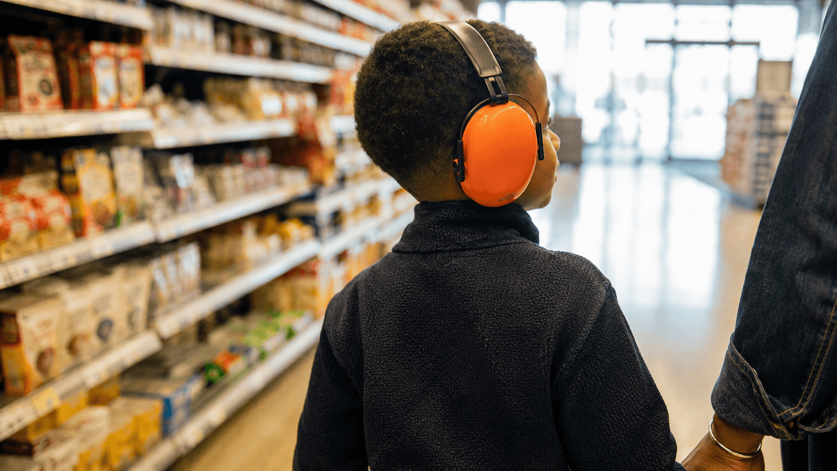 Young boy wearing orange ear defenders walks calmly down a supermarket aisle holding his parent's hand