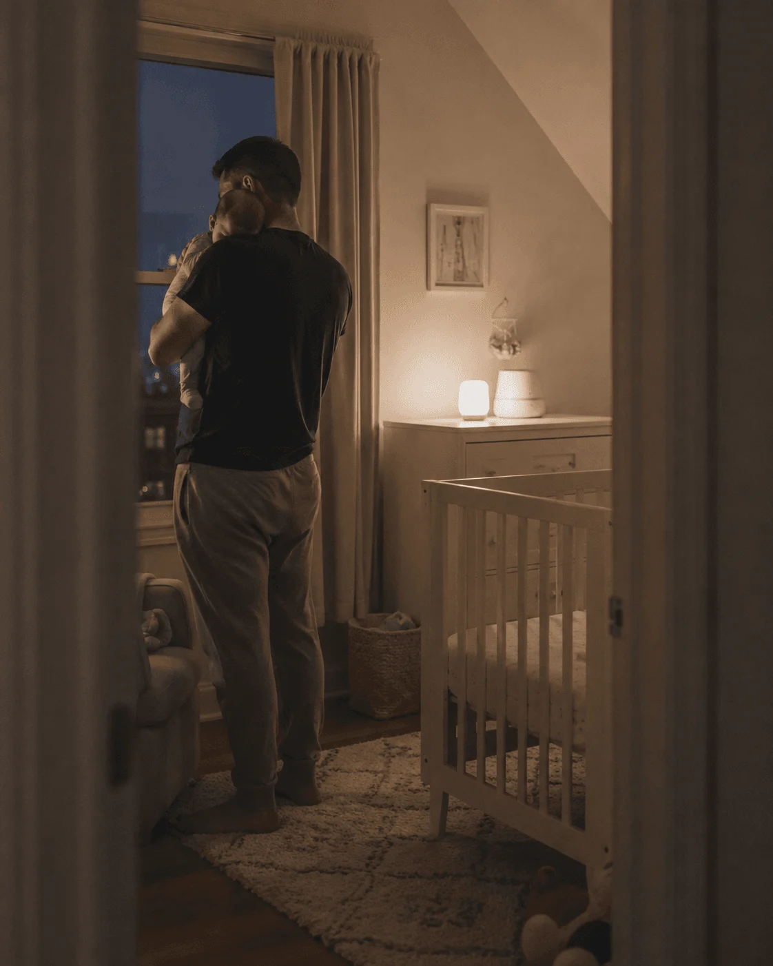 Father holds his sleeping baby at a nursery window at night, small lamp glowing beside the crib