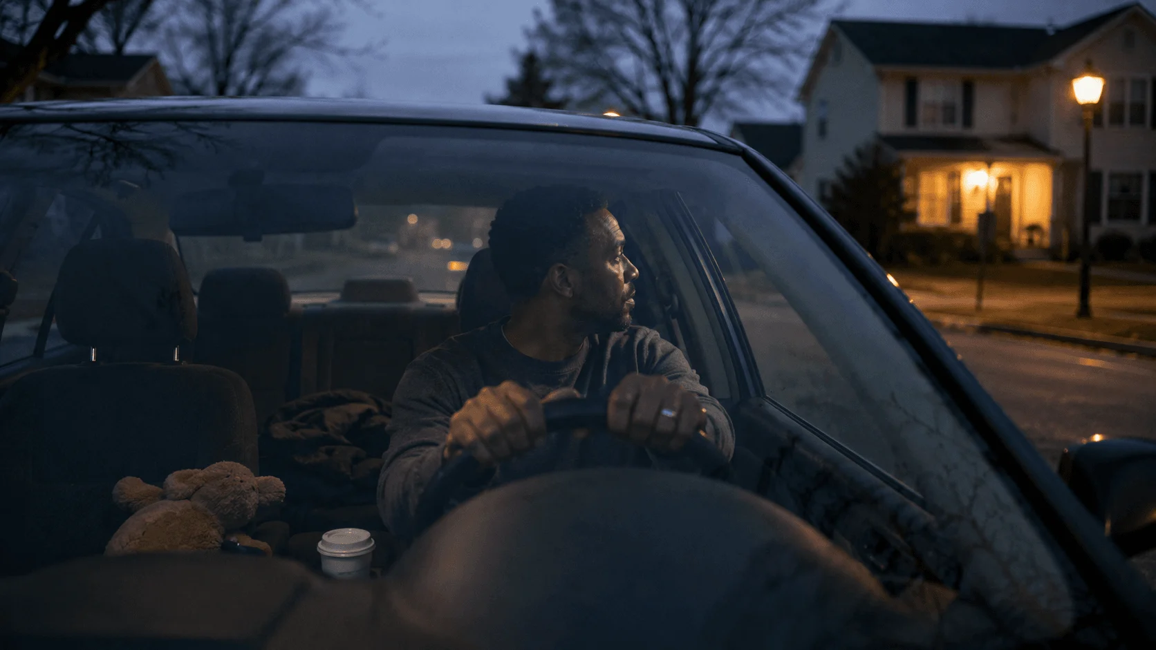 Father sits alone in his parked car at dusk, hands on the wheel, warm house lights down the street
