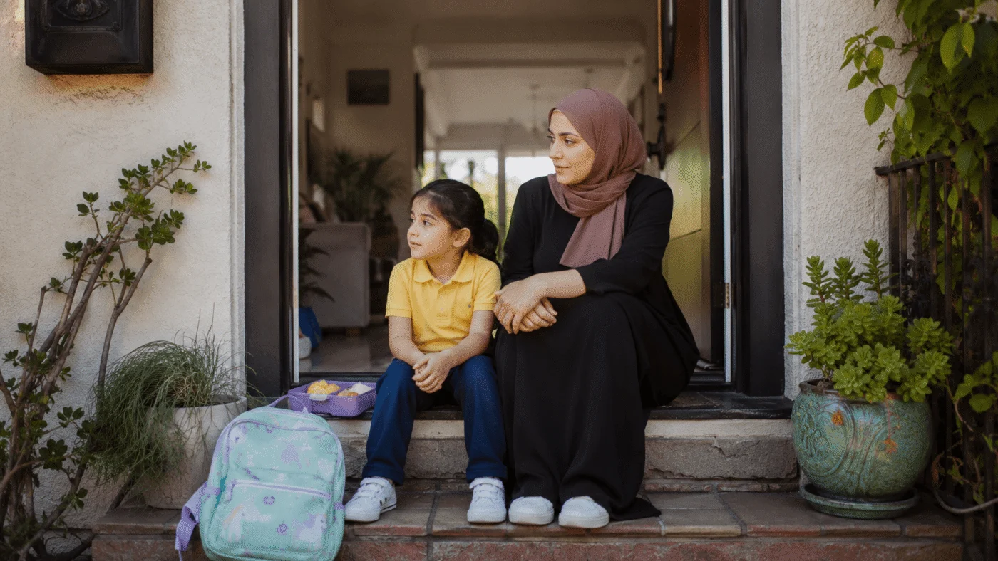 A mother in a hijab and her young daughter sit together on their front doorstep after school, backpack and open lunchbox on the step below them