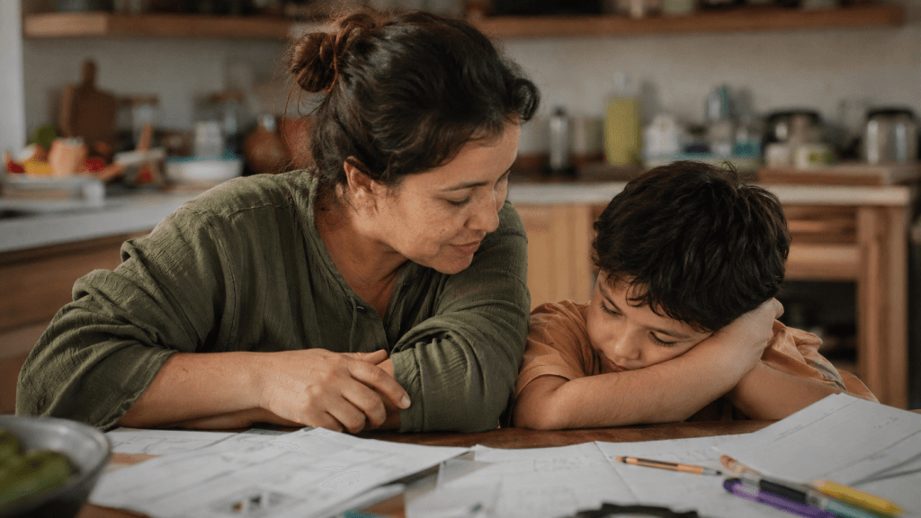 A mother leans in close beside her young son at a homework-covered kitchen table as he rests his cheek on his arm