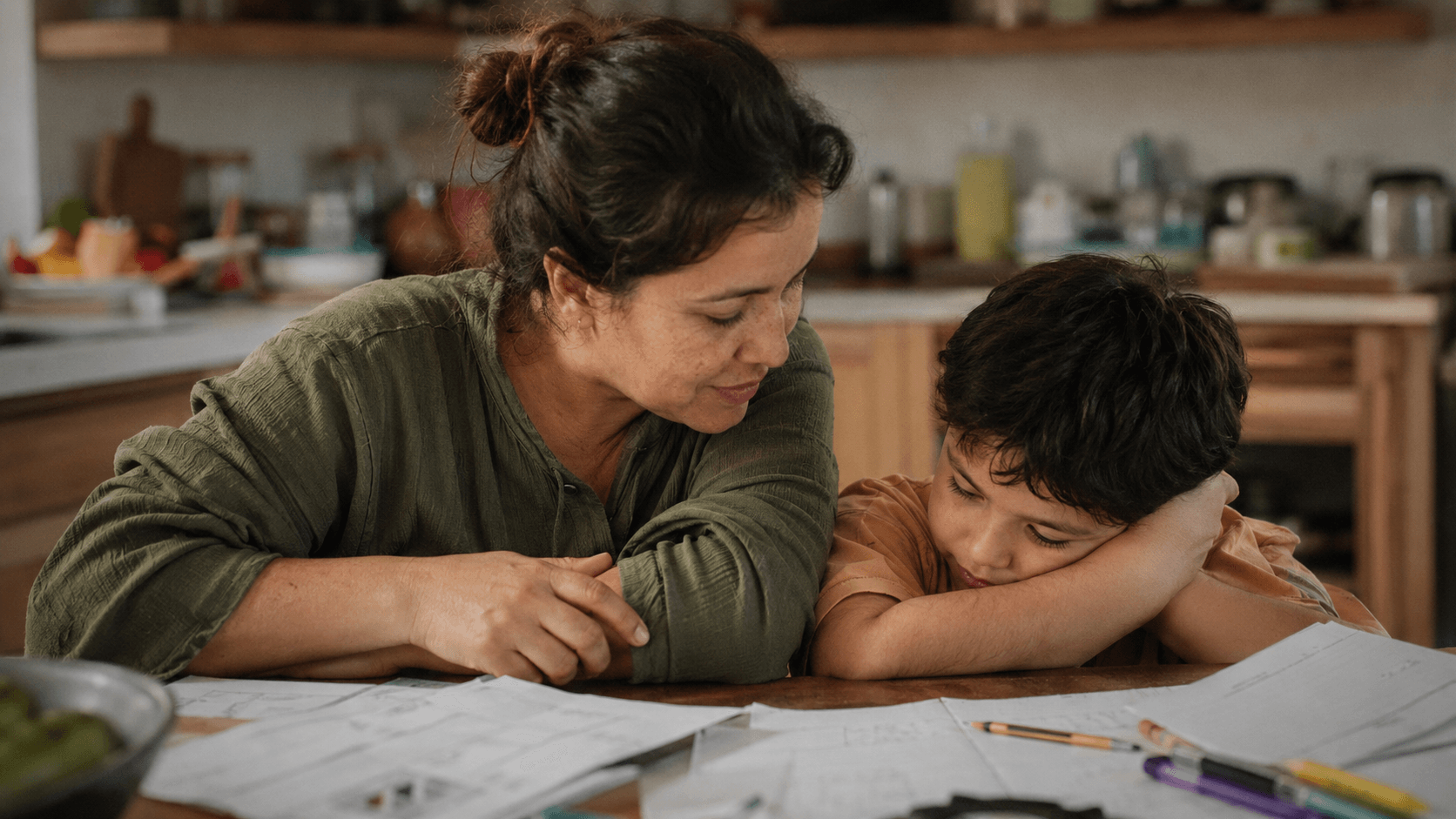 A mother leans in close beside her young son at a homework-covered kitchen table as he rests his cheek on his arm