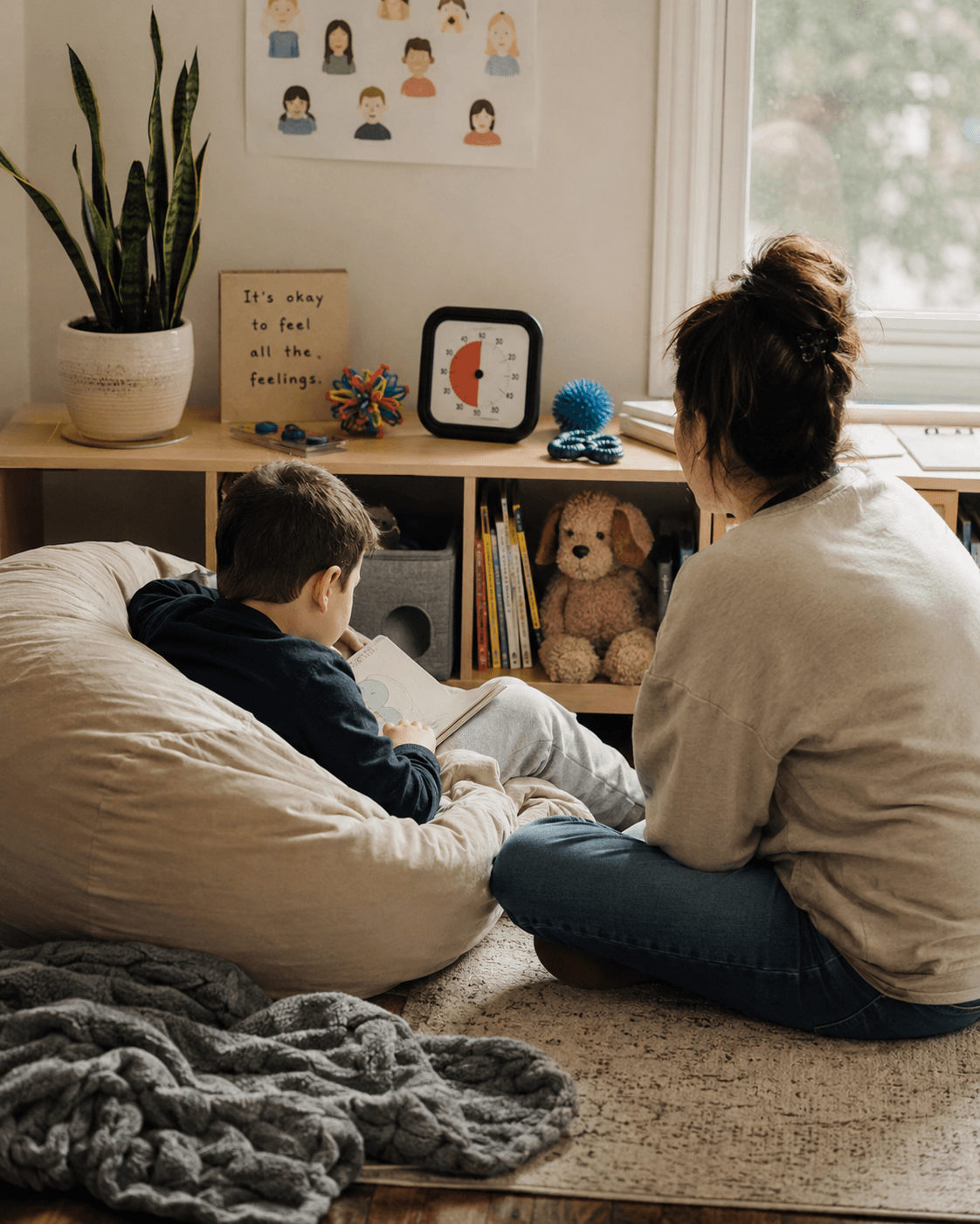 A mother and son sit together in a calm corner reading a book from the shelf while a feelings poster hangs on the wall behind them, unnoticed